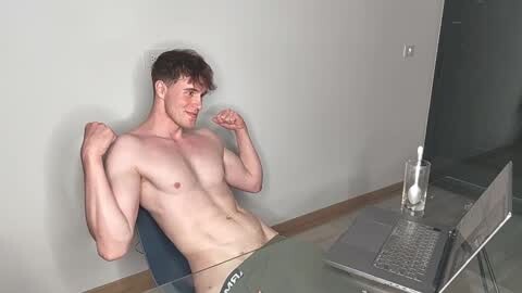 Media: Video of a muscular, light-skinned, young man with short brown hair, shirtless, flexing his biceps while sitting at a desk with a laptop and a glass of milk.