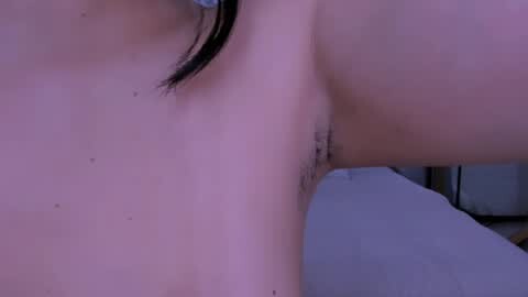 Media: Video of a close-up view of a woman's armpit, showing natural hair growth and light skin tone. The background is blurred, with a hint of a white surface and a dark object.