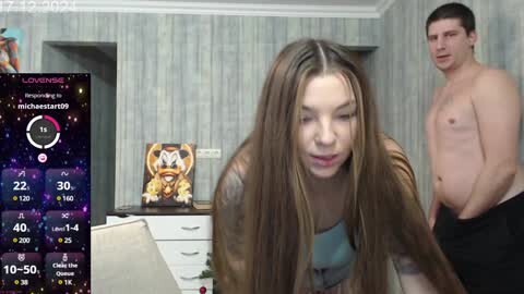 Media: A photo of a young woman with long brown hair, wearing a grey top, and a shirtless man in a room with light grey wallpaper and a TV screen displaying a video game.