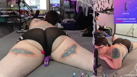 Media: Video of a plus-size woman with tattoos, lying face down on a bed, wearing black sheer shorts and a tank top, with a purple vibrator inserted.