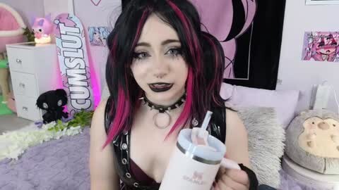 Media: Video of a pale-skinned, black-haired woman with pink highlights, wearing a black choker and leather top, holding a pink and white drink. Background includes posters, plush toys, and a pink wall.