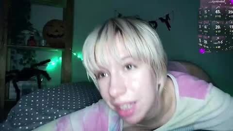 A video of a young woman with short, platinum blonde hair, wearing a striped shirt, lying on a bed, smiling and looking directly at the camera. The room is dimly lit with green fairy lights and a Halloween-themed wall calendar.