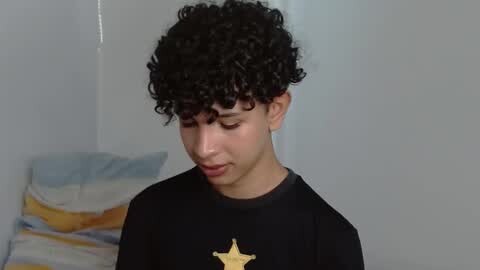 Media: Video of a young boy with curly dark hair, wearing a black shirt with a gold star emblem, looking downward in a simple room with light-colored walls and a bed with blue and white sheets.