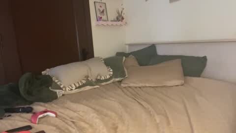Media: Video of a messy, unmade bed with beige sheets, green pillows, and a dark green comforter. A pink pair of socks is on the floor.