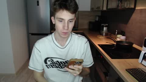 Media: Video of a young Caucasian male with short brown hair, wearing a gray T-shirt with a graphic design, holding a smartphone, in a modern kitchen with wooden countertops and stainless steel appliances.