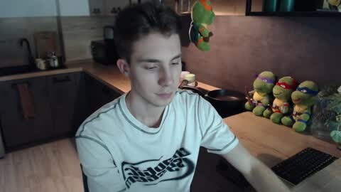 Media: Video of a young, fair-skinned, short-haired boy with closed eyes, wearing a white T-shirt, sitting at a wooden desk in a modern kitchen. Ninja Turtle toys hang above.