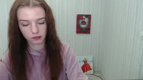 Media: A video of a young, fair-skinned woman with long, straight brown hair, wearing a light pink turtleneck, standing in front of a wall with a red painting and a framed video of a woman.