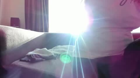Media: A video captures a person's upper torso and arm, wearing a white t-shirt and glasses, lying on a bed with a gray blanket. The room has a bright, sunlit window with a white curtain. The image is slightly blurry with lens flares.