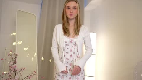 Media: Video of a young, slim, fair-skinned woman with long, straight blonde hair, wearing a white floral dress with a white cardigan, standing in a softly lit, modern room with white walls and a tall, narrow mirror.