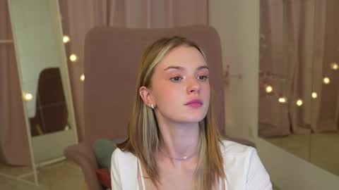 Media: Video of a young woman with fair skin, straight blonde hair, and a neutral expression, wearing a white top. She sits in a room with pale pink curtains and glowing string lights.