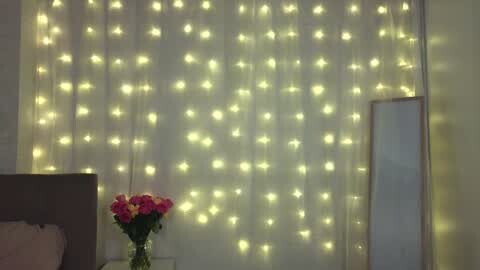 Media: Video of a cozy bedroom with a wall of glowing, star-shaped fairy lights, a vase of pink and yellow flowers on a nightstand, and a standing mirror to the right.