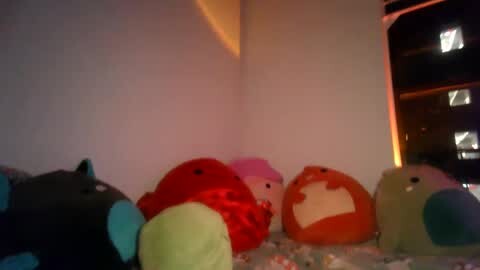 Media: Video of a cozy, dimly-lit room with a soft, orange plush toy and a colorful, polka-dotted blanket on a bed. The background features a corner with a dark window and a warm, amber light.