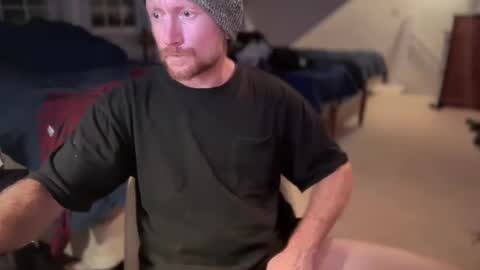 Media: A video of a man with a beard, wearing a grey beanie and black T-shirt, sitting on a couch in a dimly lit room with beige carpet.