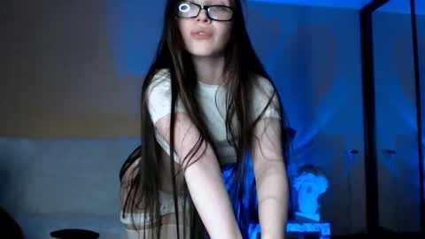 Media: Video of a young woman with long dark hair, glasses, and a white top, bending over a bed in a dimly lit room with blue lighting.