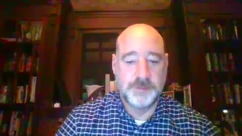 Media: A video of a bald, bearded man with a serious expression, wearing a plaid shirt, sitting in a dimly lit room with wooden bookshelves filled with books.