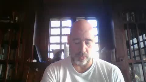 Media: Video of a bald, bearded white man in a white shirt, sitting indoors with wooden bookshelves and a window with a grid pattern in the background.