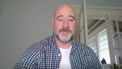Media: Video of a bald, middle-aged white man with a mustache and beard, wearing a blue and white checkered shirt, standing indoors with white blinds and furniture in the background.