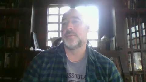 Media: Video of a middle-aged man with a beard, wearing a green plaid shirt over a grey T-shirt, sitting indoors in a dimly lit room with wooden bookshelves, a window, and a TV in the background.