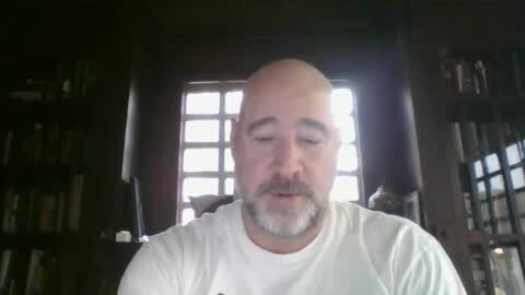 Media: Video of a middle-aged, bald, white man with a trimmed beard, wearing a white shirt, sitting indoors with dark wooden bookshelves and a large window in the background.