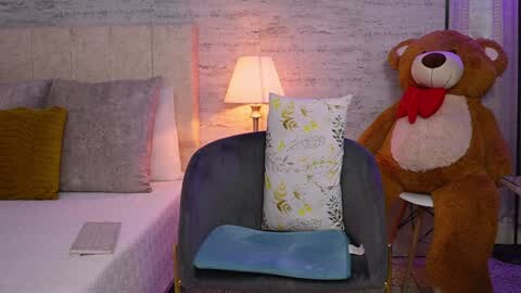 Media: A cozy, dimly-lit bedroom scene with a plush, grey armchair featuring a light blue cushion and a white pillow with green floral print. A large teddy bear and a yellow cushion sit on the left. The background includes a white bed with gray pillows, a soft yellow throw blanket, and a table lamp emitting warm light.