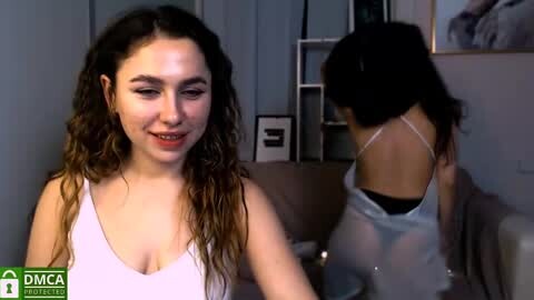 Media: Video of a young woman with curly hair in a light pink tank top, smiling, in a living room with a woman in a white bra and black underwear visible in the background.