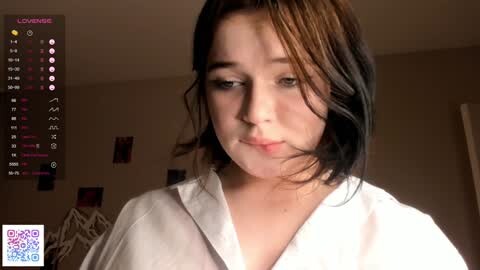 Media: Video of a young woman with shoulder-length dark hair, wearing a white shirt, indoors with a beige wall and decorative items in the background.