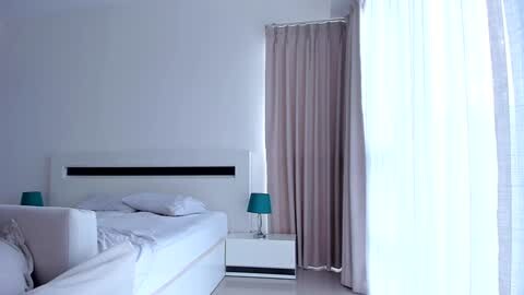 Media: Video of a minimalist, modern bedroom with a white bed, white curtains, and beige drapes. The room features a teal lampshade and a plain white wall.