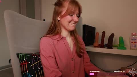 Media: Video of a smiling red-haired woman in a pink blouse, seated in a modern living room with a colorful plaid cushion and a shelf of vibrators.