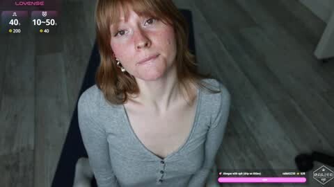 Media: A video of a Caucasian woman with red hair and freckles, wearing a grey cardigan, kneeling on a blue mat in a dimly lit room.