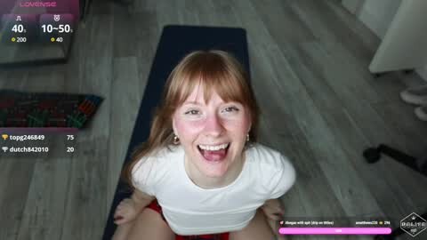 Media: Video of a smiling, red-haired woman on a yoga mat, wearing a white t-shirt, with a blue keyboard and wooden floor in the background.