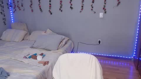 Video of a minimalist bedroom with a beige wall, a white bed, a plush white chair, and a blue LED strip lighting the ceiling. The bed is unmade, with a few scattered toys and a stuffed animal.