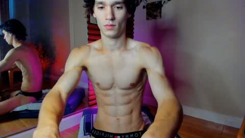 Media: Video of a shirtless, muscular young man with short, dark hair, wearing black Calvin Klein underwear, sitting on a bed in a dimly lit room with purple lighting. Another person, partially visible, is also on the bed.