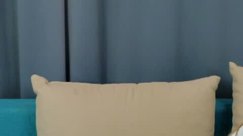 Media: Video of a beige pillow on a teal couch, with a dark blue curtain background.