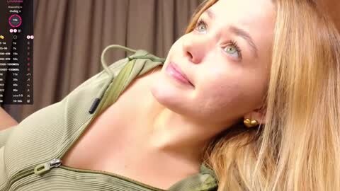 Media: Video of a fair-skinned, blonde-haired woman with blue eyes, wearing a green ribbed top, lying on her side, looking upward. Background features a brown curtain.