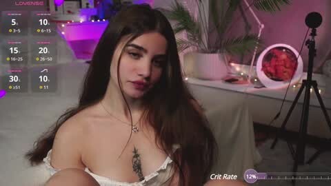 Media: A video of a young woman with long, dark hair, wearing a white off-shoulder top, sitting on a bed with a large round mirror on a tripod in the background.