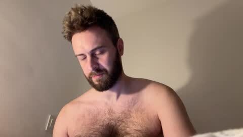 Media: Video of a bearded, light-skinned man with curly hair, appearing relaxed and shirtless, against a plain beige wall, creating a casual, intimate atmosphere.