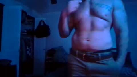 Media: Video of a shirtless man with a muscular build, wearing tan pants, in a dimly lit room with a blue hue. He stands in front of a closet with a black handbag hanging inside.