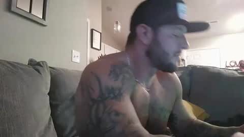 Media: Video of a shirtless man with tattoos, wearing a black cap, sitting on a grey couch in a dimly lit living room.