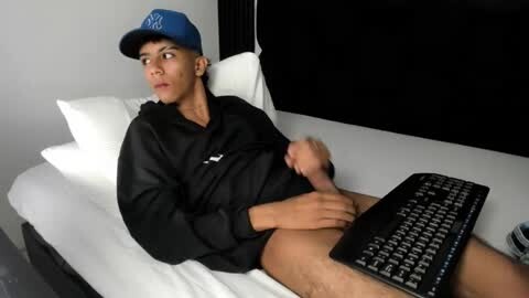 Media: Video of a young man in a black hoodie and blue baseball cap, lying on a bed with white sheets, holding a black keyboard, looking thoughtful.
