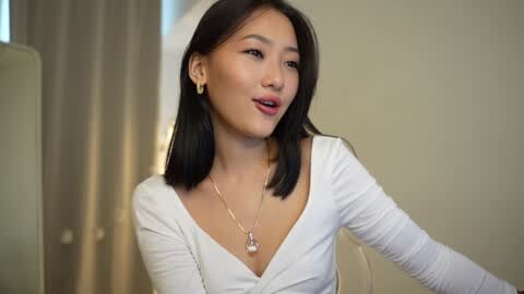 Media: Video of a young Asian woman with straight black hair, fair skin, and slender physique, wearing a white long-sleeve top, smiling softly, in a modern, minimalist indoor setting.