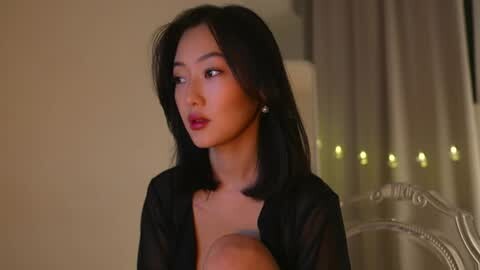 Media: Video of an Asian woman with shoulder-length black hair, wearing a black sheer top, standing indoors, looking contemplative.