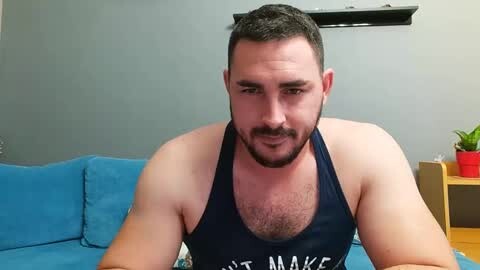 Media: Video of a muscular, bearded man with short dark hair in a navy tank top, seated on a blue couch against a gray wall.