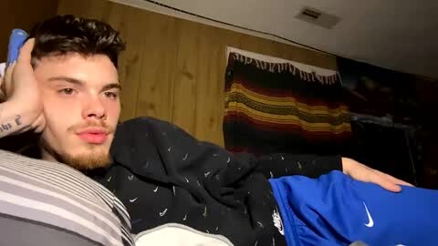 Media: Video of a young man with short brown hair, light skin, and a beard, lying on a bed in a dimly lit room. He wears a blue Nike shirt and blue shorts. A colorful woven blanket hangs on the wall behind him.