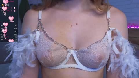 Media: Video of a fair-skinned woman wearing a sheer, white lace bra with feathered shoulder straps, highlighting her medium-sized breasts. Background includes a dimly lit room with soft purple lighting and a blurred, green plant.