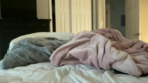 Media: Video of a messy bedroom with a rumpled white bedspread, a light pink blanket, and a dark blue blanket. A black dresser and white double doors are visible in the background.