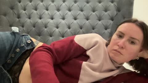 Media: A video of a young woman with pale skin, dark hair, and a neutral expression, lying on a grey tufted headboard in a room with a beige wall. She wears a maroon and white sweater and blue jeans.