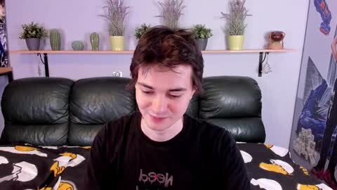 Media: A video of a young man with short brown hair, wearing a black shirt with a yellow duck pattern, smiling while sitting on a green leather couch in a room with a lavender wall and potted cacti.