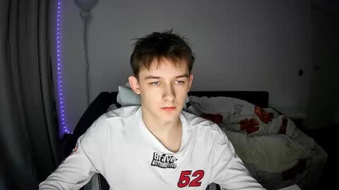 Media: Video of a young Caucasian man with short brown hair, wearing a white \"Beavers\" jersey, sitting on a bed with a floral-patterned blanket in a dimly lit room.