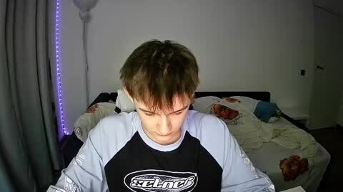 Media: Video of a young boy with short, light brown hair, wearing a black and white \"Rip Curl\" hoodie, sitting on a bed with a white comforter featuring a floral pattern. The room has dim lighting and a string of blue LEDs on the wall.