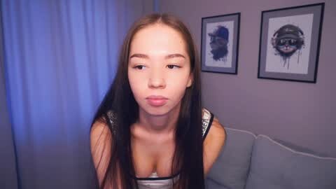 Media: Video of a young Asian woman with long black hair, wearing a black tank top, puckering her lips in a pout, in a modern, minimalist room with gray sofa and abstract art on the wall.